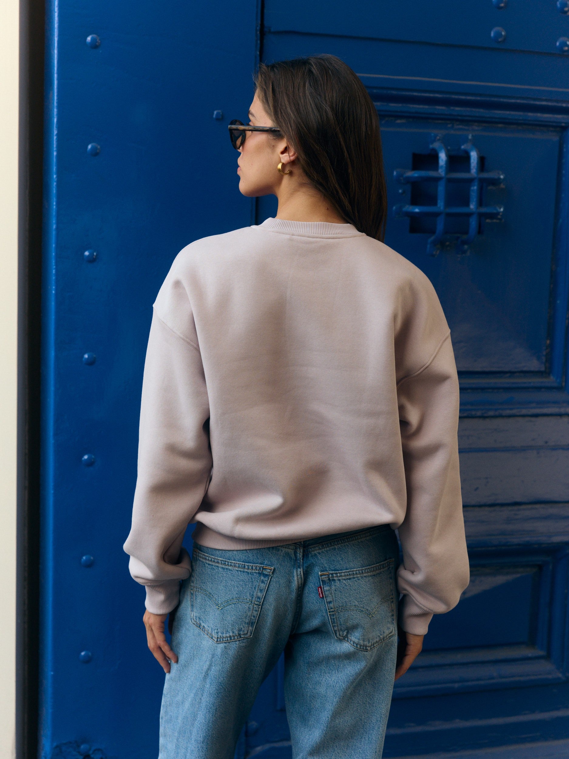 Back view of a woman wearing a dusty pink crewneck sweatshirt against a vibrant blue door.